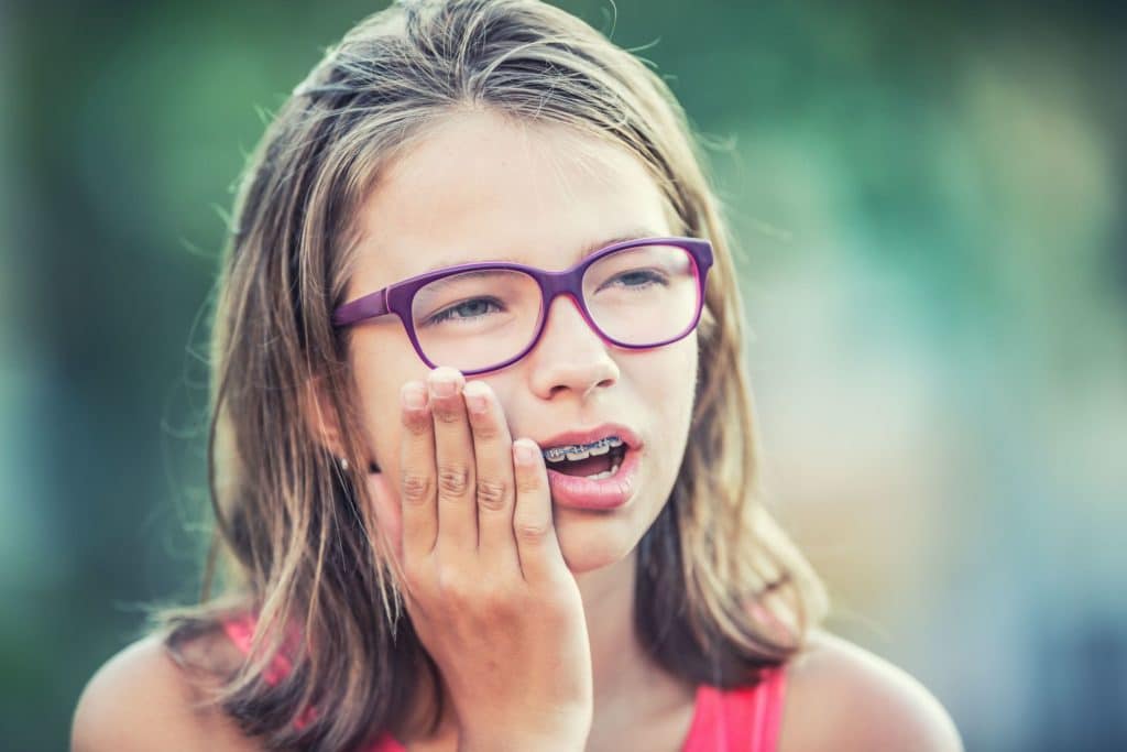 Preteen girl with metal braces and purple glasses applying pressure to sore gums