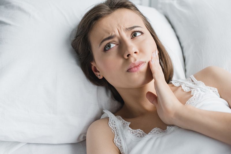 Worried young woman with tootache holding side of mouth in bed